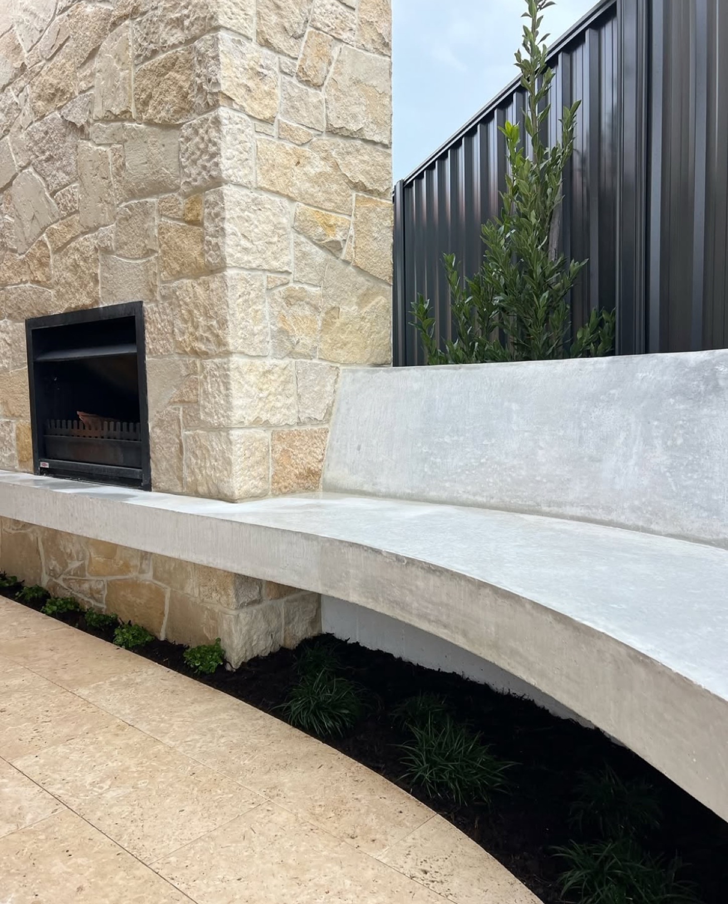 Curved concrete bench seat sweeping past a stone-clad outdoor fireplace, garden bed tucked underneath