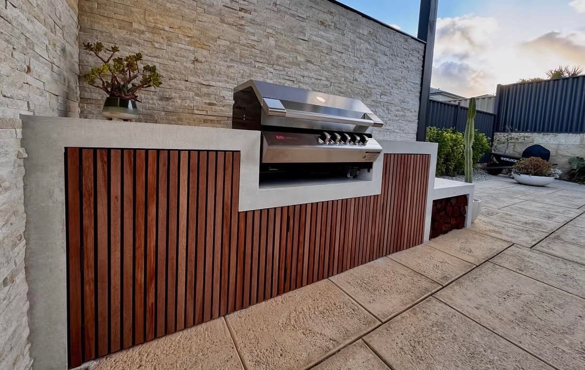 Outdoor kitchen with a honed concrete benchtop and timber slat front, built-in grill set into the run and a stacked stone wall behind