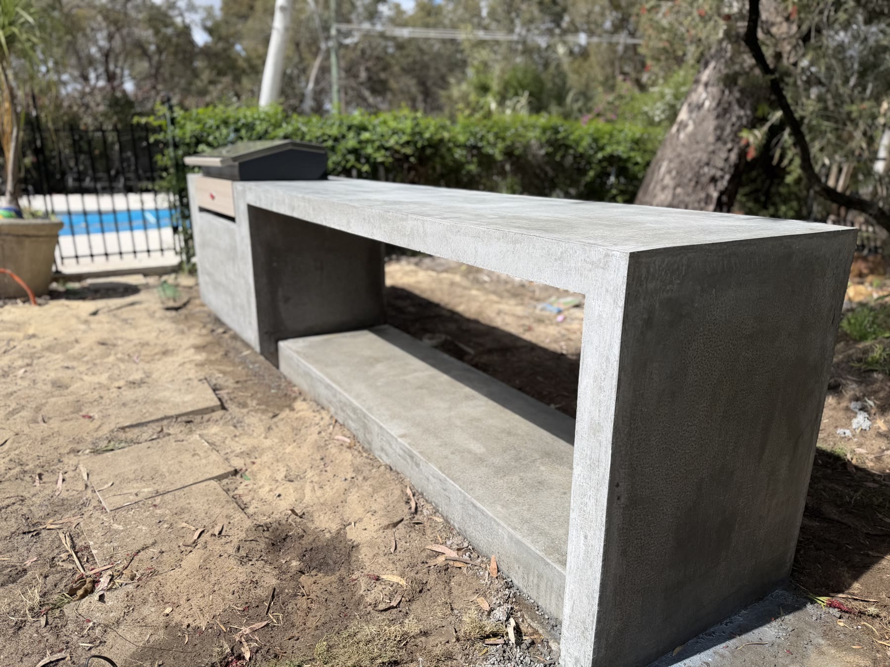 Architectural grey burnish BBQ bench in Kalamunda from the side, cantilevered shelf over the poured base with the pool behind